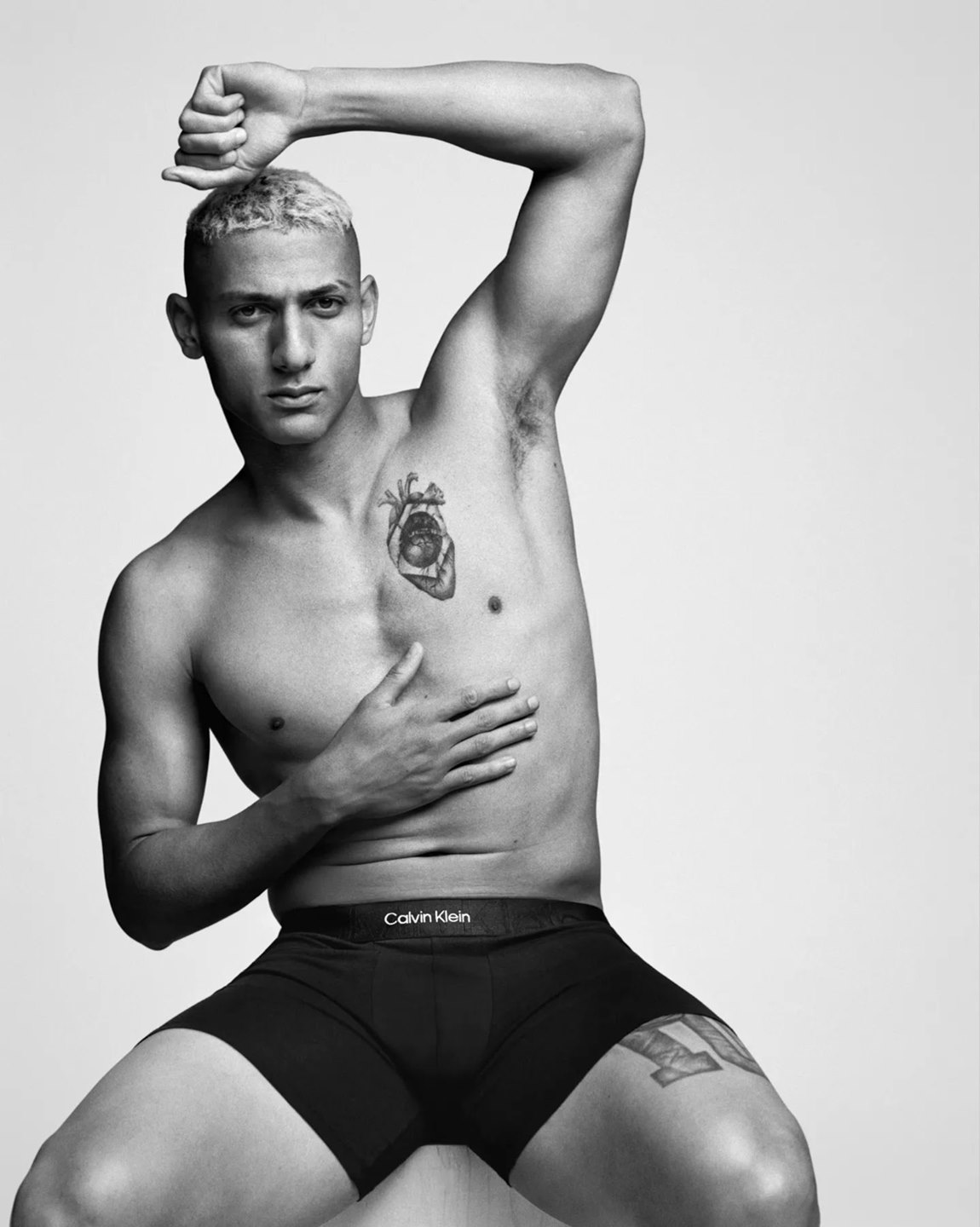 O jogador Richarlison, um homem jovem, branco, com cabelo crespo raspado e descolorido de loiro, posa em um fundo branco liso. A foto é uma campanha da Calvin Klein. Ele usa uma cueca preta da marca Calvin Klein. - Metrópoles