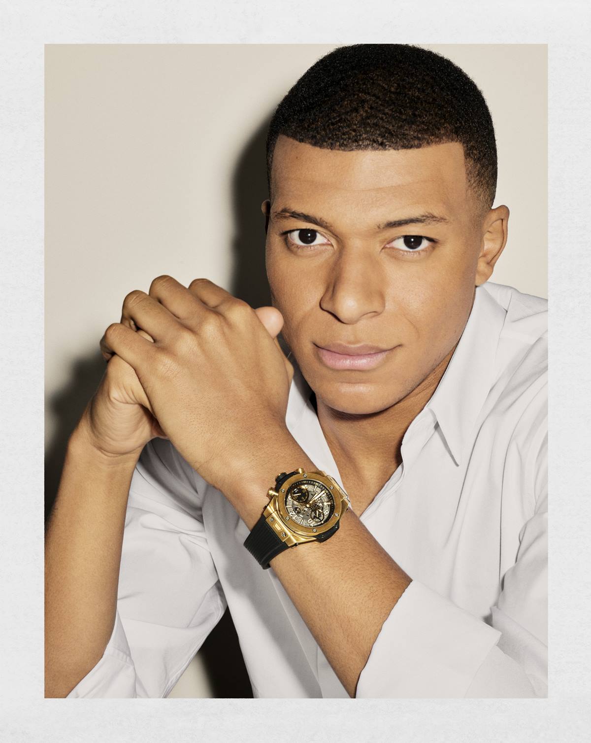 O jogador Kylian Mbappé em foto de campanha da marca de relógios de luxo Hublot. Ele usa uma camisa de botão branca e um relógio preto com detalhes de ouro. - Metrópoles