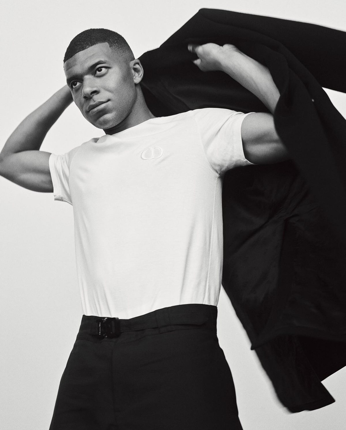 O jogador Kylian Mbappé em ensaio de fotos para a revista Wall Street Journal. Posa em um fundo branco liso. Usa uma camiseta branca lisa, uma calça preta de alfaiataria e um blazer preto. - Metrópoles