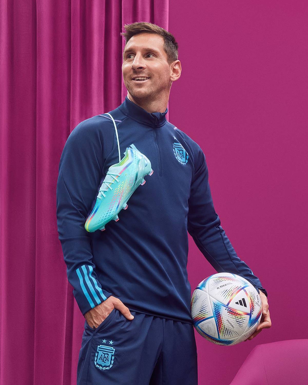 O jogador Lionel Messi, um homem jovem e branco, de cabelo liso, posando para foto publicitária da marca esportiva Adidas. Usa um conjunto de casaco e bermuda, ambos azul escuro com detalhes azuis claro, e está com uma chuteira pendurada nos ombros. Segura uma bola de futebol na mão esquerda. - Metrópoles