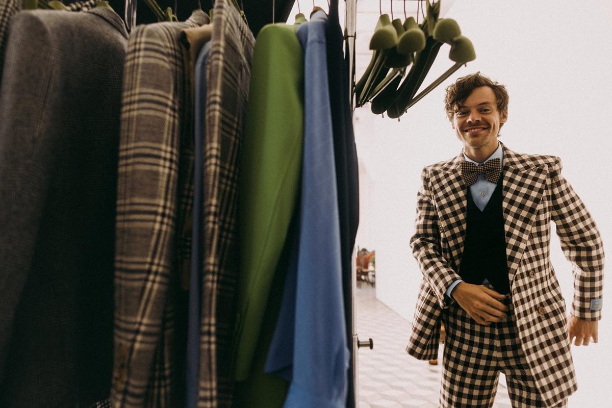 O cantor Harry Styles, um homem jovem, branco, com cabelo liso castanho, nos bastidores de uma sessão de fotos com a marca Gucci. Ele usa uma camisa azul, um pulôver cinza, gravata e um conjunto de calça e blazer xadrez. Todos os itens são parte de uma colaboração entre Harry Styles e a Gucci. - Metrópoles