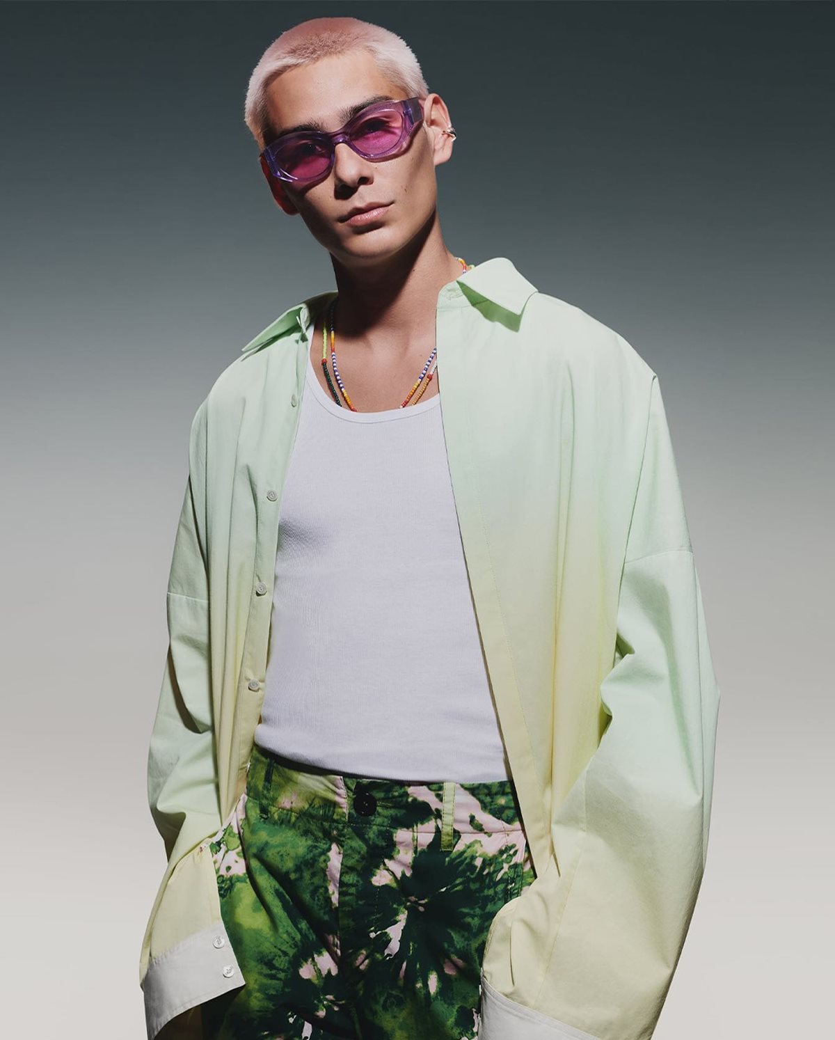 Homem asiático, jovem e com cabelo liso curto e descolorido, posa para foto em um fundo branco. Ele usa uma regata branca, uma camisa verde clara e um calça verde escura com estampa de plantas. A foto é de uma campanha publicitária da marca Zara. - Metrópoles