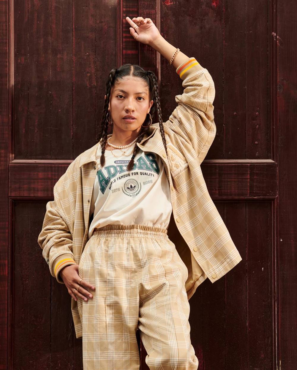 Uma mulher negra e jovem, de cabelo trançado castanho, posa para foto em frente a uma porta de madeira. Ela usa um top branco e um conjunto de calça e blazer xadrez marrom. As peças e a foto são da campanha publicitária da marca Adidas. - Metrópoles