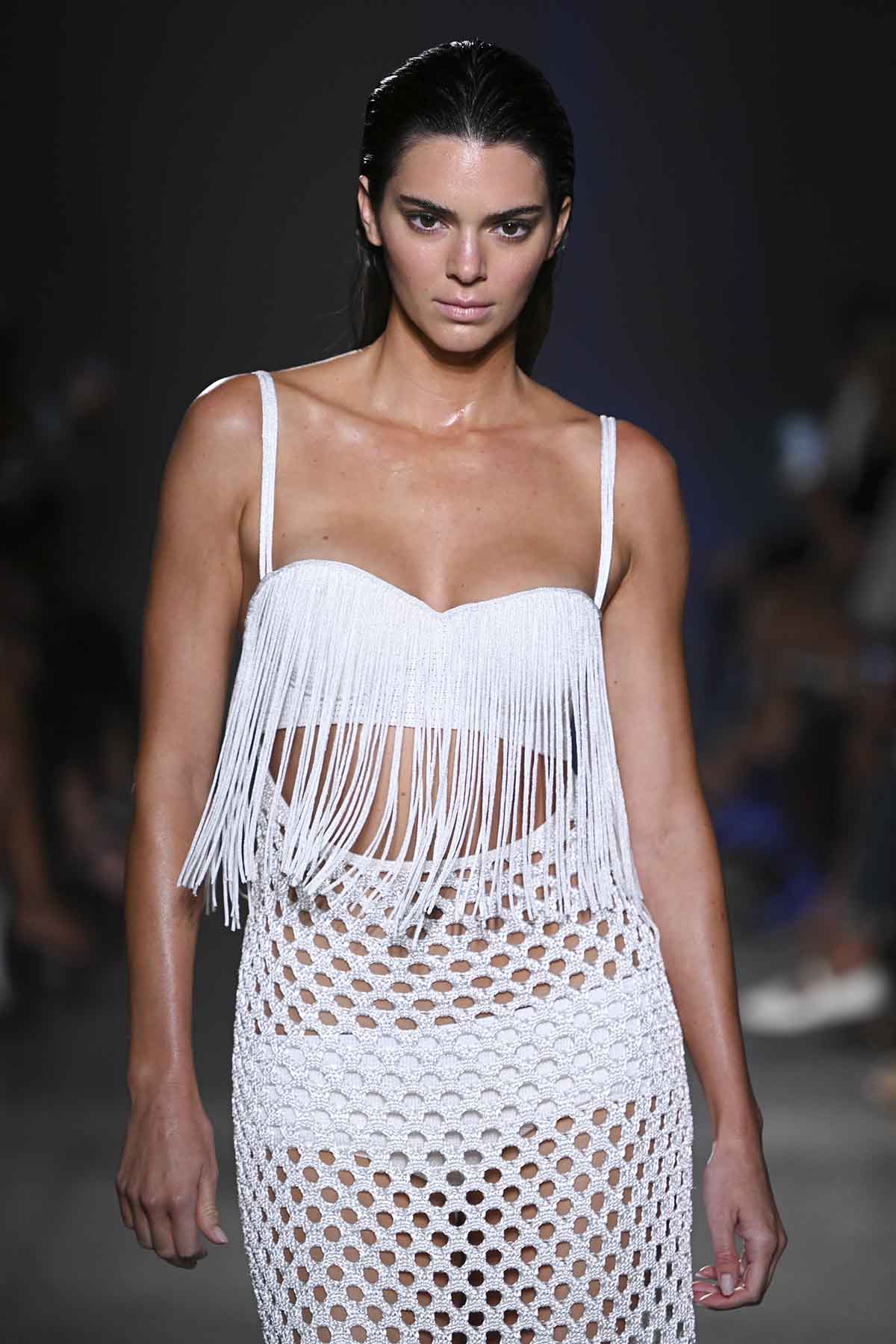 A modelo Kendall Jenner, uma mulher branca e jovem, com cabelo castanho liso, desfilando na Semana de Moda de Nova York em setembro de 2022. Usa um conjunto branco de top e saia da marca Proenza Schouler. O top possui um babado de franjas na parte de baixo. - Metrópoles