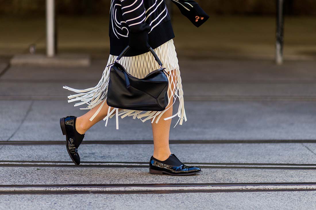 Foto de uma mulher branca e jovem andando pela rua. Na imagem é possível ver apenas da sua cintura para baixo. Ela usa um vestido branco com franjas, um casaco de moletom preto, um sapato estilo mocassim de couro preto e uma bolsa, também de couro e preta. - Metrópoles