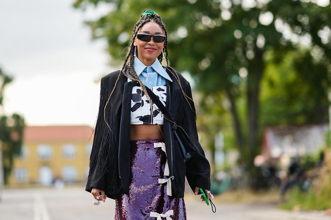 Mulher negra e jovem, de cabelos trançados longos, posando para foto durante a Semana de Moda de Copenhagem. Ela usa uma camisa azul de botão curta, um top preto e branco curto com estampa de vaca, uma jaqueta preta com franjas, uma saia de paetê marrom e um óculos escuros preto.