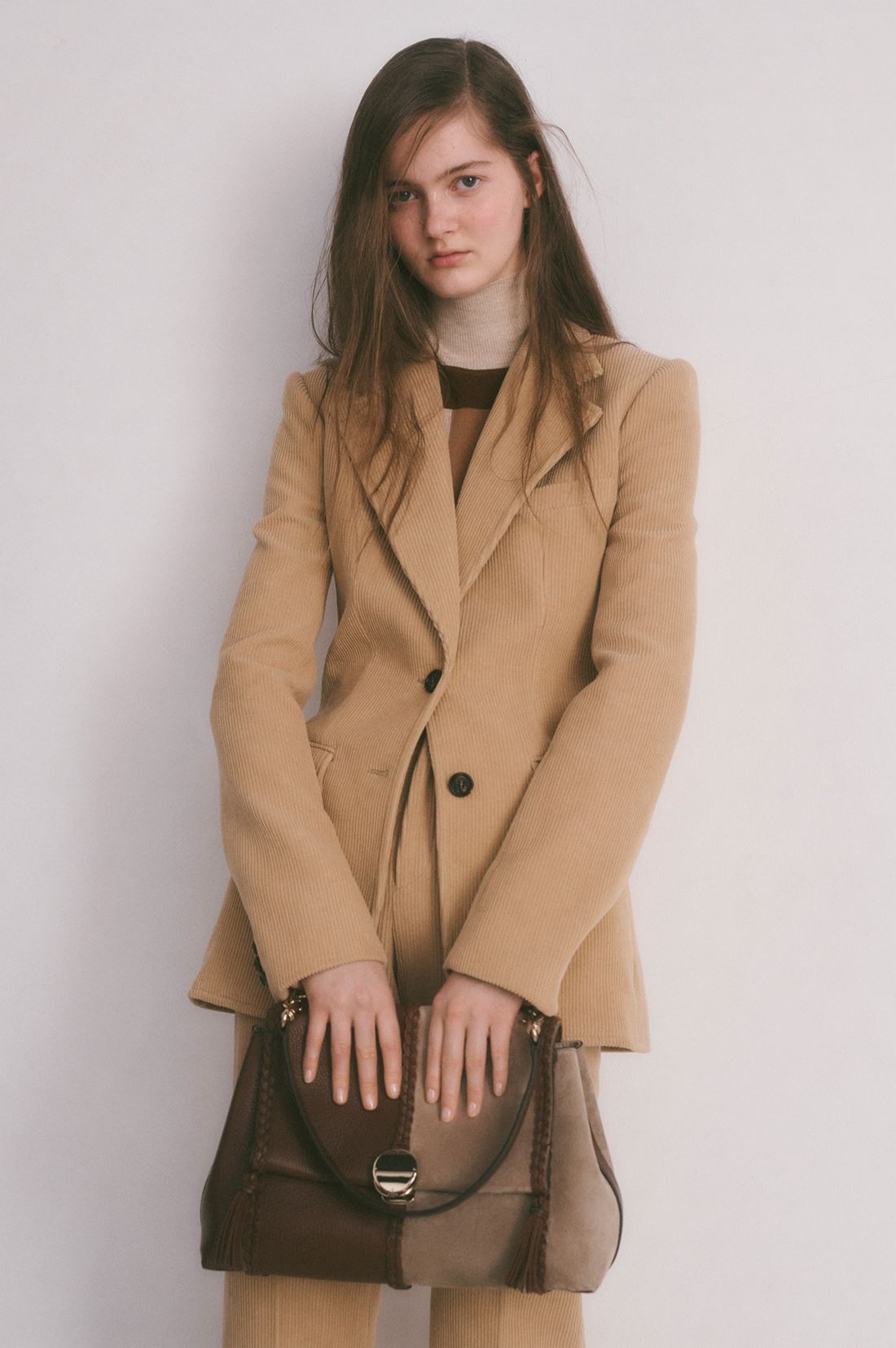 Modelo branca e jovem, com cabelo liso e loiro longo, posando para foto em um fundo branco. A foto é de uma campanha da marca Chloé.. Ela usa uma camiseta marrom e um conjunto de calça e blazer, ambos de camurça bege. - Metrópoles