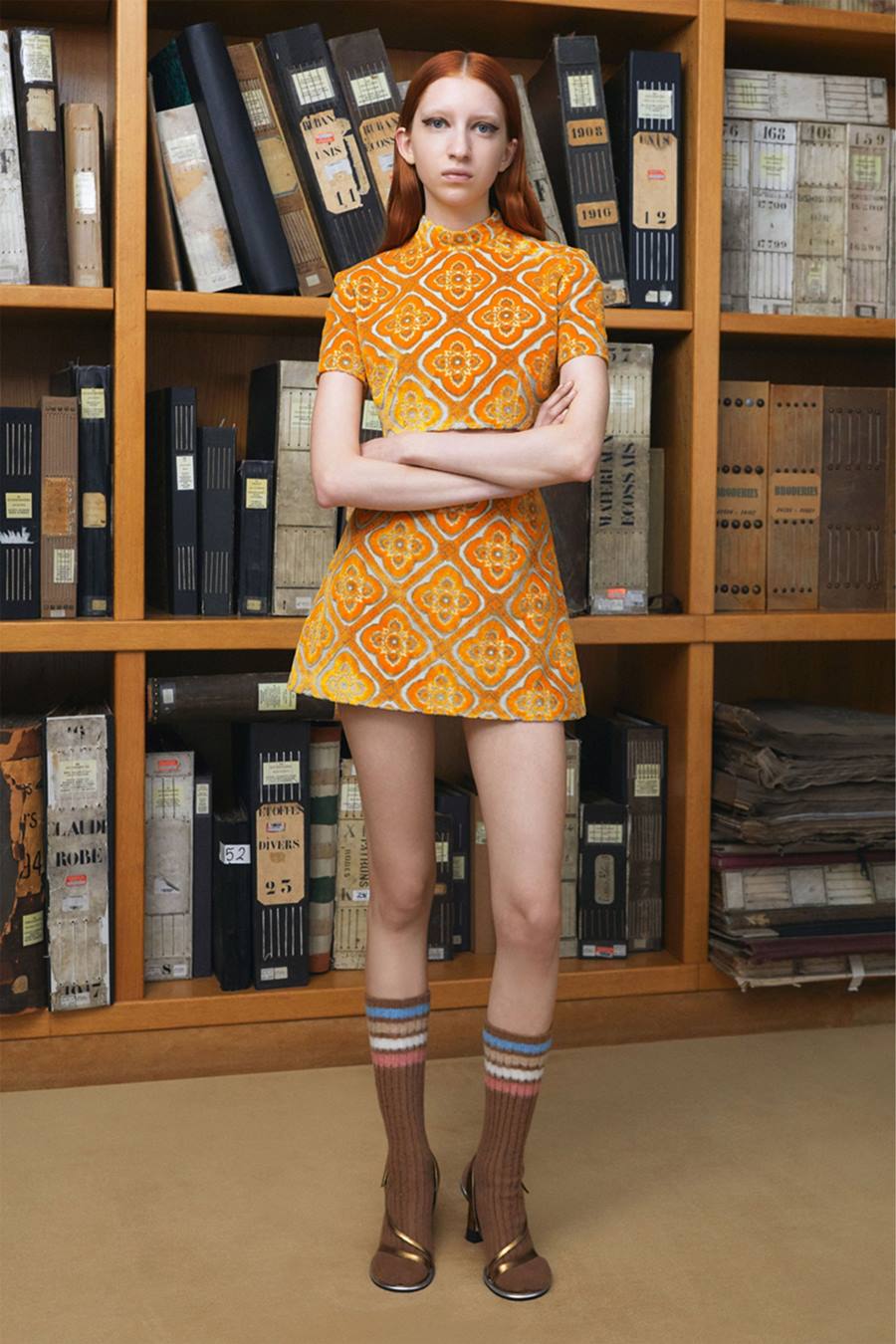 Modelo branca e jovem, com cabelo liso e ruivo longo, posando para foto em frente a uma estante de livros. A foto é de uma campanha da marca Etro. Ela usa conjunto de top cropped e saia laranja, meias 3/4 e salto alto. - Metrópoles