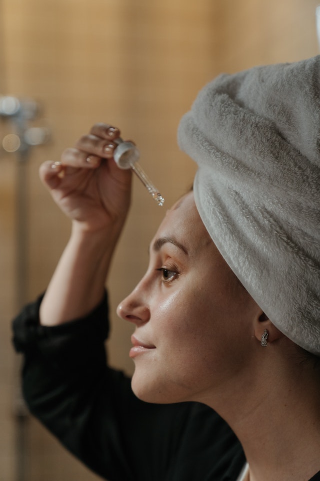 Mulher com toalha no cabelo de frente para o espelho aplicando produto no rosto - Metrópoles