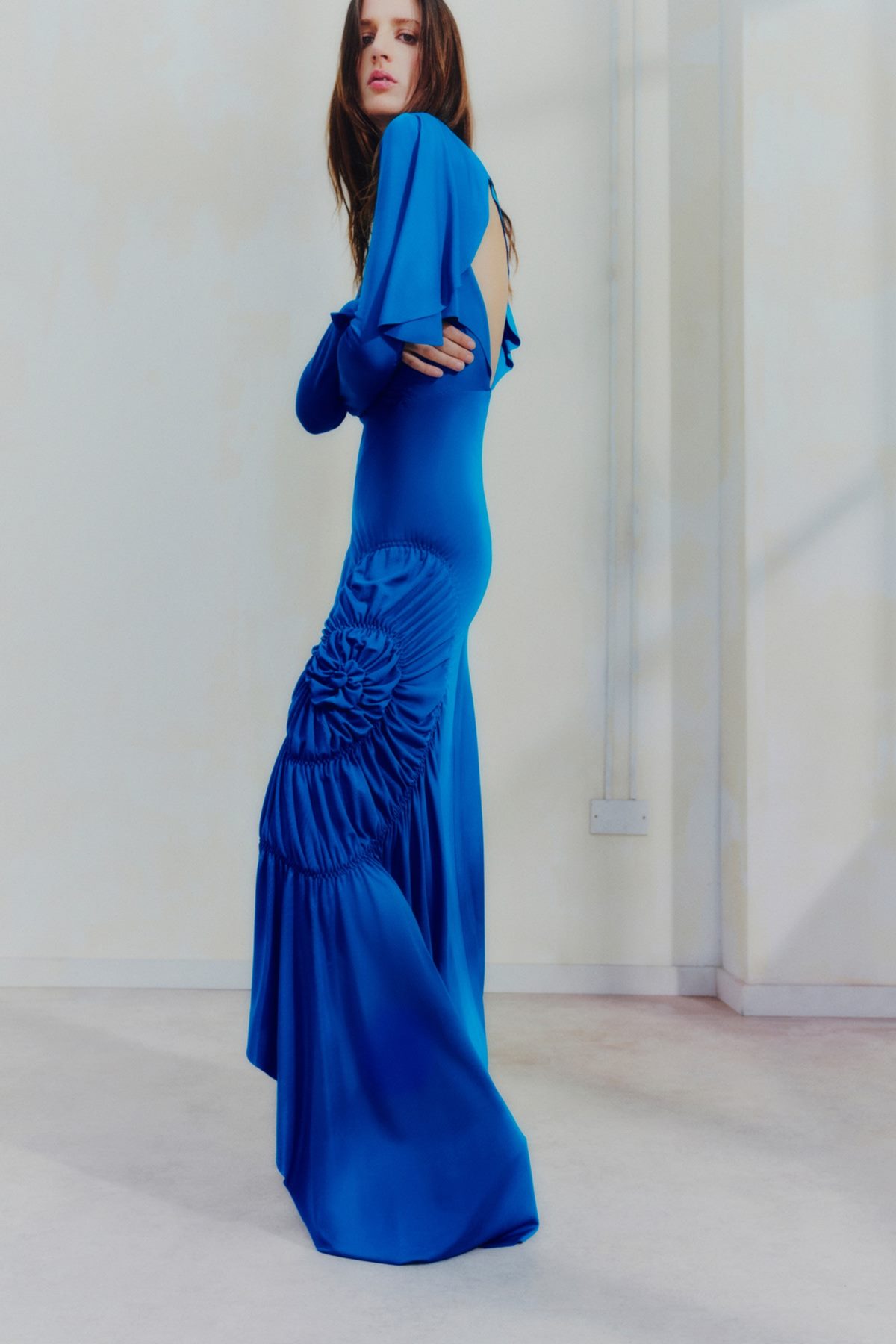 Foto de campanha da marca Victoria Beckham. Na imagem é possível ver uma mulher branca e jovem, de cabelo liso longa, com um vestido longo azul turquesa. - Metrópoles