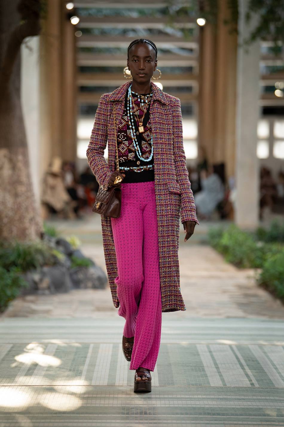 Modelo negra e jovem, com cabelo trançado, andando na passarela do desfile Pre-Fall 2023 da marca Chanel. Ela usa uma calça roxa folgada, uma camiseta preta e um casaco rosa e preto por cima. - Metrópoles