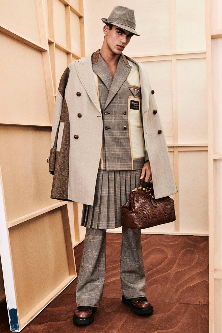 Homem moreno e jovem, de cabelo curto castanho, em foto de campanha de divulgação da marca Moschino. Ele usa um conjunto de blazer, saia e calça, todos de alfaiataria e na cor cinza. Por cima, usa um casaco trench coat bege com detalhes em marrom. - Metrópoles