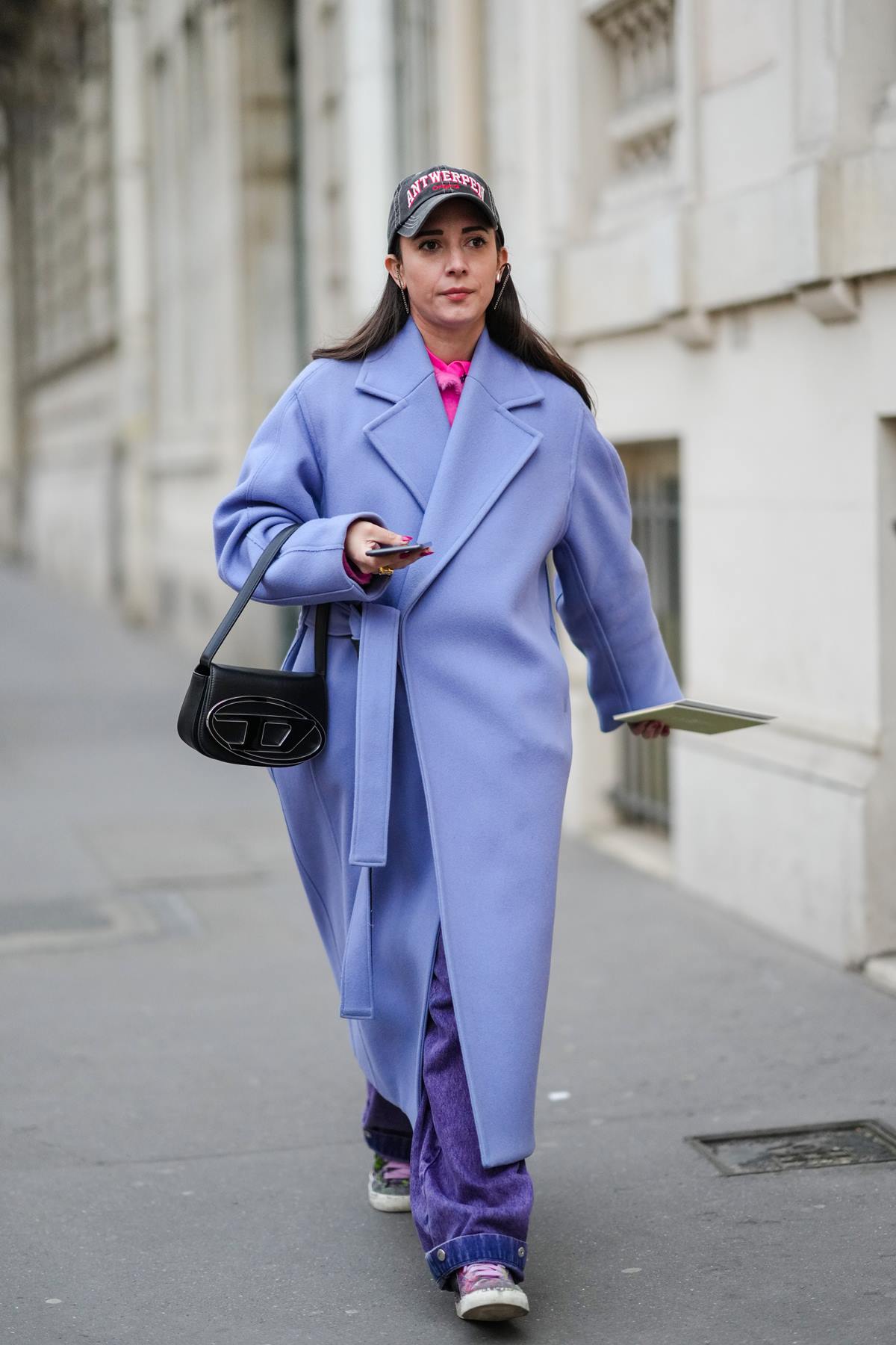 Uma mulher branca e jovem, de cabelo liso longo, anda pela rua durante a Semana de Moda de Paris. Ela usa uma camisa rosa, uma calça jeans folgada e, por cima, um casaco lilás de inverno. Segura com o braço direito uma bolsa preta de couro da marca Diesel chama 1DR. - Metrópoles