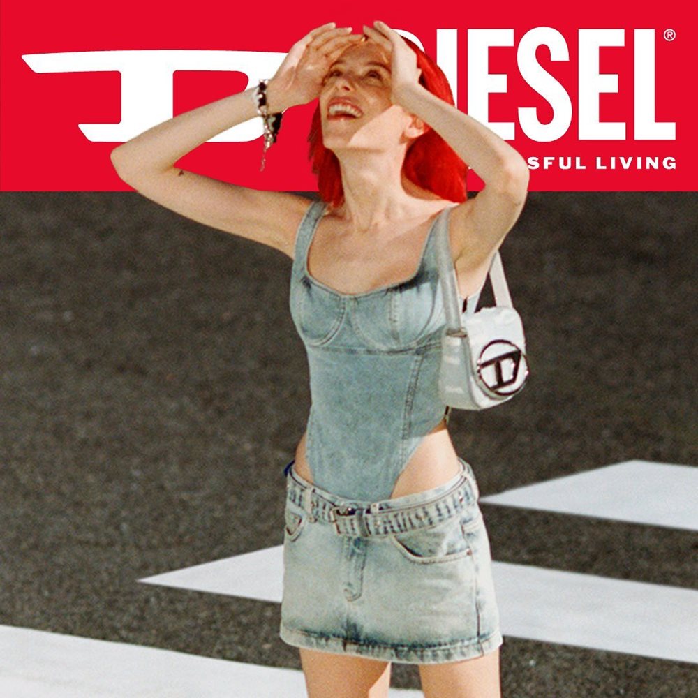 Mulher branca e jovem, de cabelo liso curto e ruivo, posa em faixa de pedestres para uma foto de campanha da marca Diesel. Ela usa um top e uma saia, tudo jeans, e uma bolsa prateada curta modelo 1DR em baixo do braço. - Metrópoles