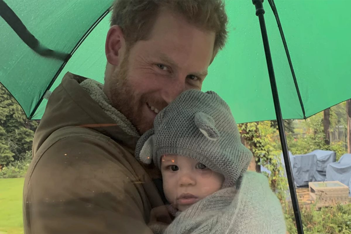 Foto colorida de homem branco e ruivo segurando guarda-chuva verde e bebê com touca de orelhinhas cinza - Metrópoles