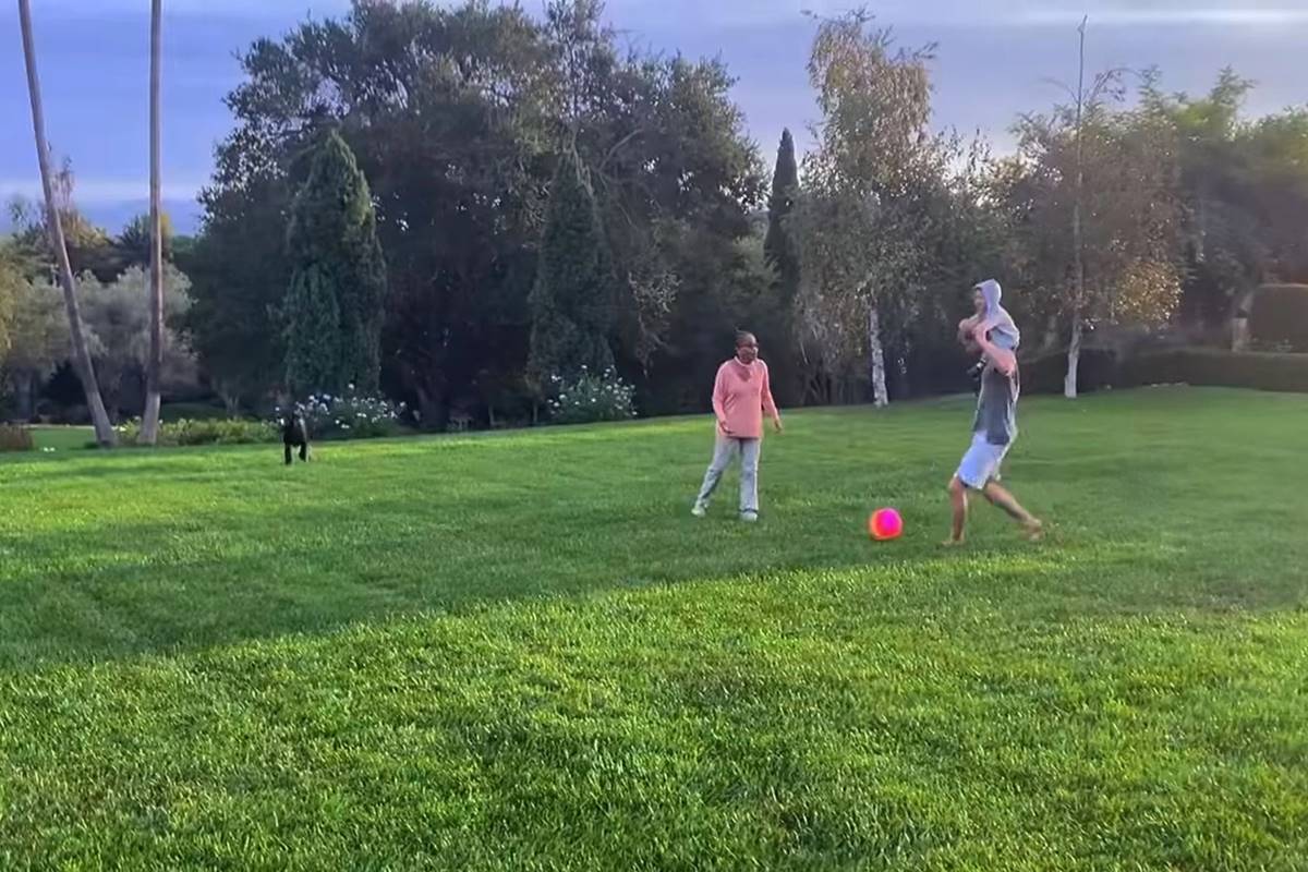 Foto colorida de mulher e homem com menino nos ombros brincando de bola em um gramado - Metrópoles