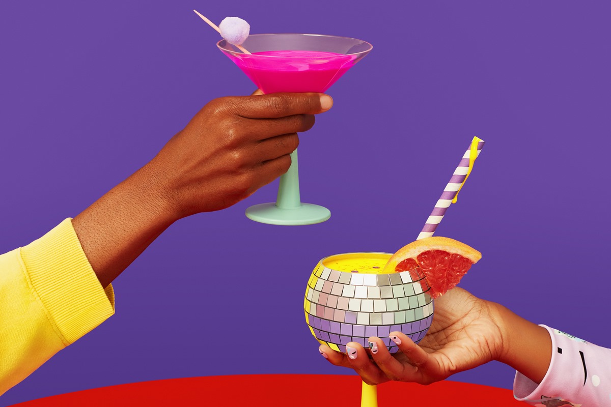 Foto feita em estúdio, com um fundo roxo, onde duas mulheres negras brindam com taças de drink. - Metrópoles