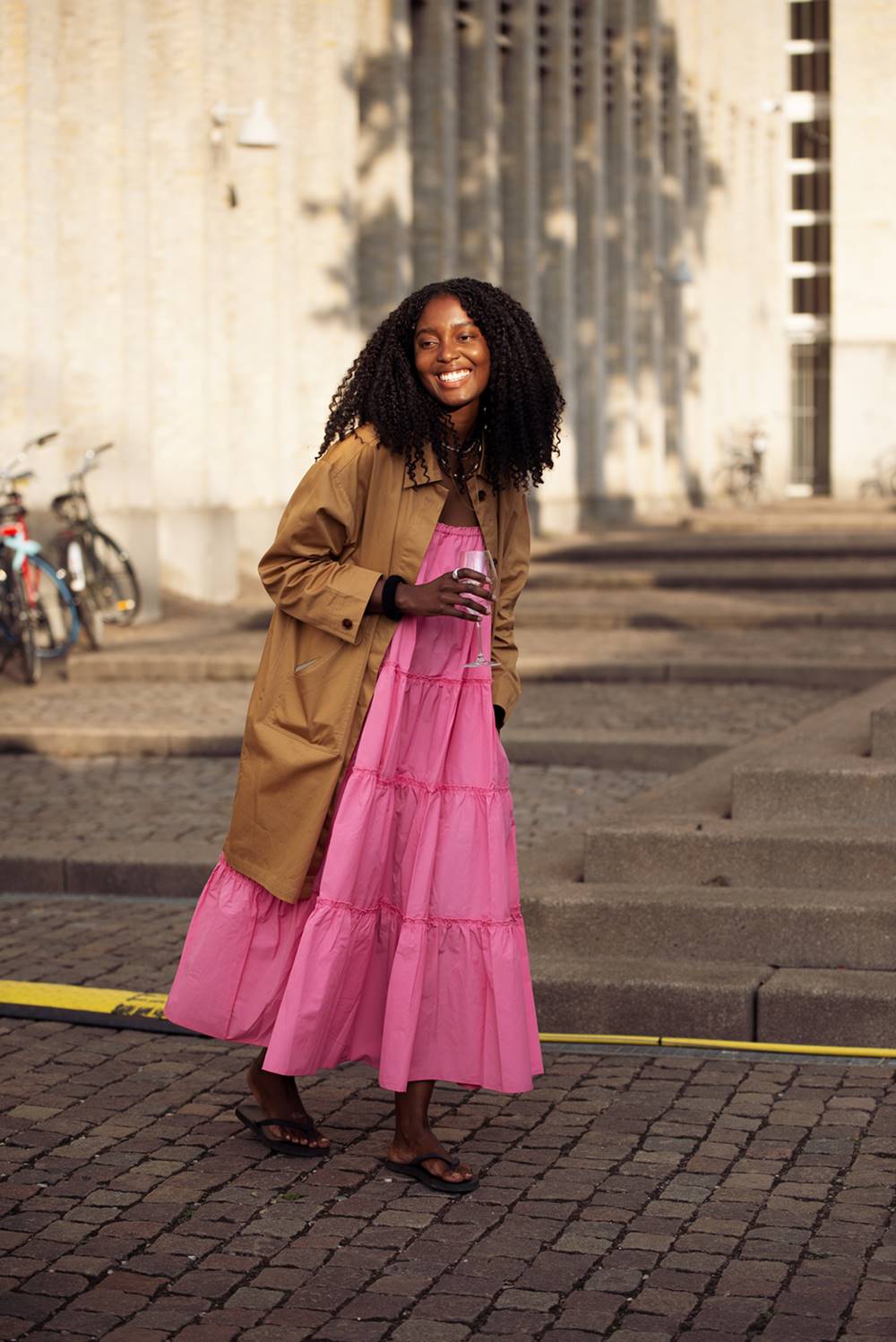 Mulher jovem e negra, de cabelo cacheado longo, posando para foto em um estacionamento. Ela usa um vestido rosa longo e folgado, um casaco bege por cima e um chinelo preto nos pés. - Metrópoles