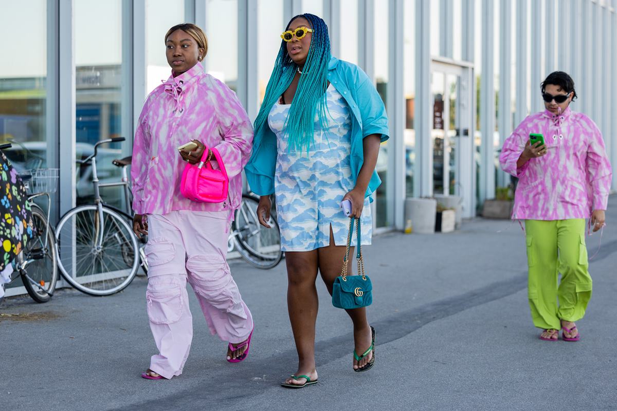 Duas mulheres jovens e negras andando pela rua durante a Semana de Moda de Copenhague. A primeira usa calça rosa e um casaco com estampa rosa e branca. A segunda usa um vestido curto com estampa azul e branca que imita o céu. Elas usam chinelos Havaians. - Metrópoles