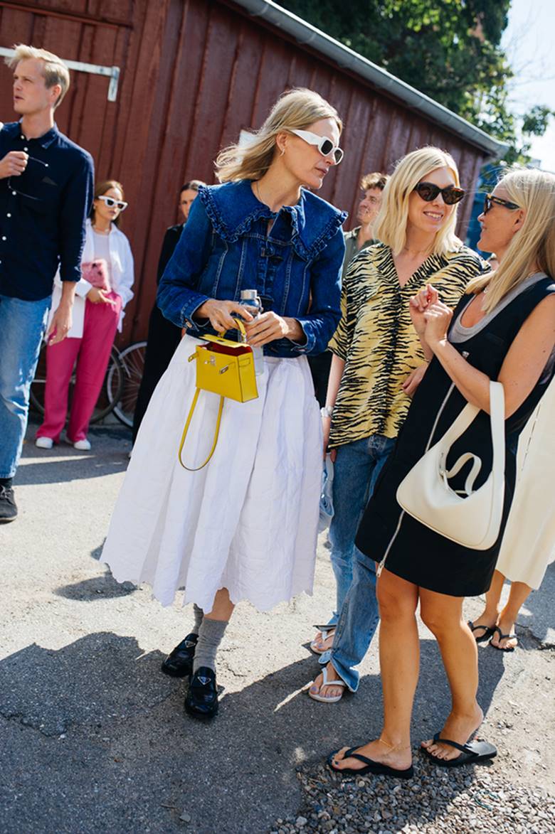 Três mulheres brancas, jovens e loiras durante a Semana de Moda de Copenhague. Elas andam pela rua. A primeira usa uma saia branca e uma blusa jeans, a segunda usa uma camisa com estampa de listras e calça jeans, a terceira usa um vestido preto. - Metrópoles