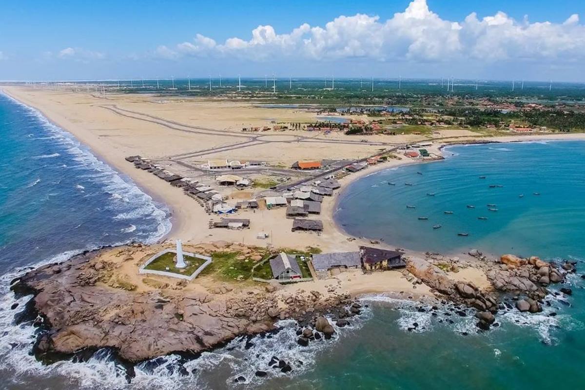 Foto colorida de praia, com pedras, um forte e estabelecimentos ao redor - Metrópoles