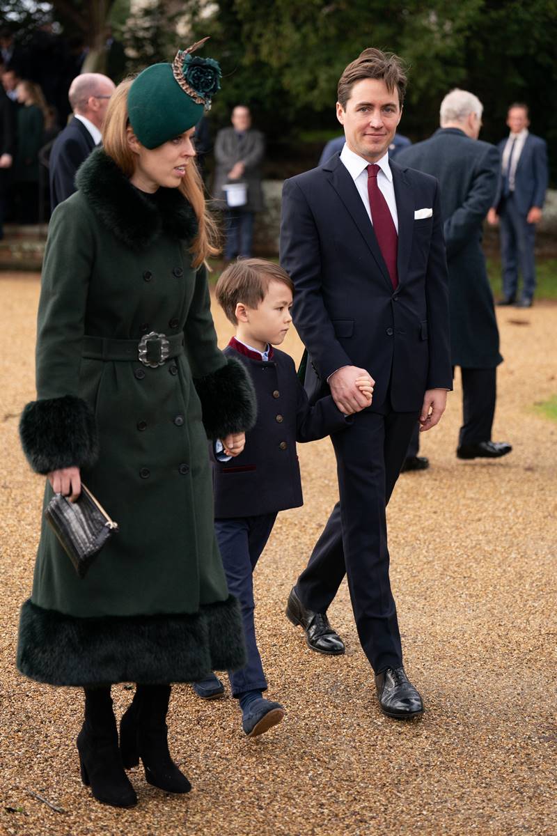 Foto colorida de mulher branca e ruiva com casaco verde de mãos dadas com menino branco e de terno de mãos dadas com homem branco vestido com terno - Metrópoles