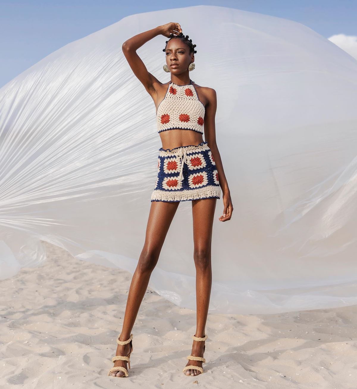 Mulher negra e jovem, de cabelo cacheado curto, posa para foto em dunas de areia. Ela usa um conjunto de top cropped e saia curta, ambas peças feitas de crochê nas cores branca, vermelha e azul. - Metrópoles