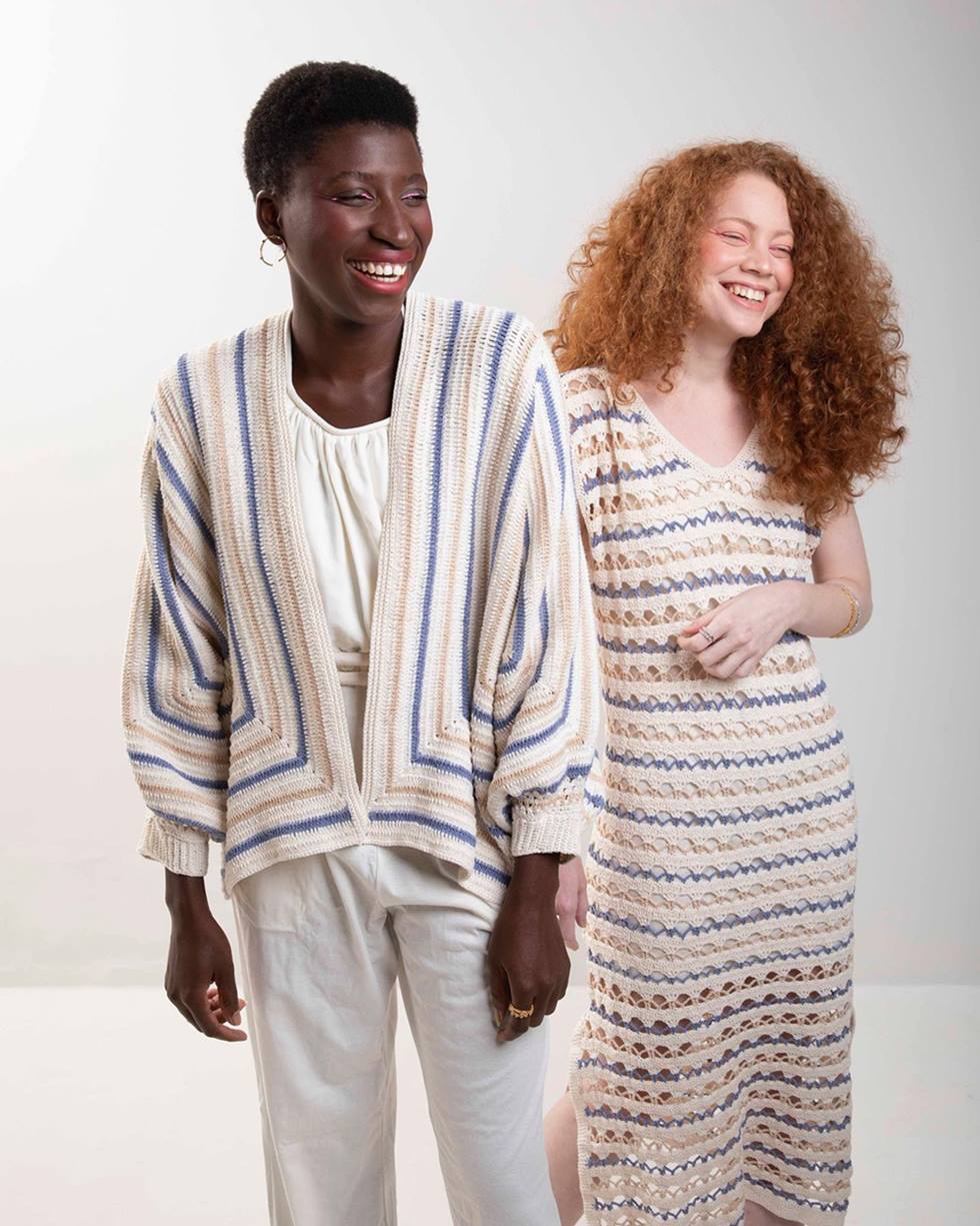 Uma mulher negra de cabelo curto cacheado e uma branca, de cabelo ruivo longo, posam para foto em um fundo branco. A primeira usa um conjunto de blusa e calça branca e um cardigan de crochê branco, azul e amarelo; a segunda usa um vestido de crochê, também branco. - Metrópoles