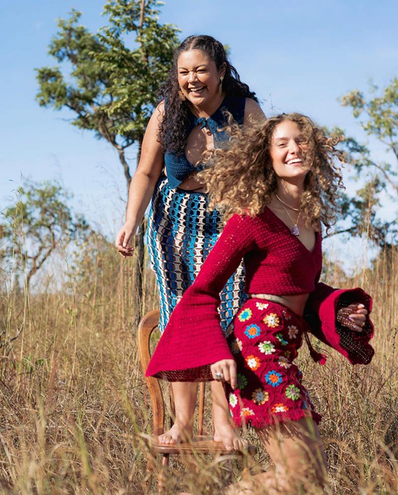 Duas mulheres brancas e jovens, uma de cabelo castanho e outra de cabelo loiro, posam para foto em meio à vegetação do cerrado. A primeira usa uma roupa azul e a segunda uma vermelha. Todas as peças são feitas de crochê. - Metrópoles