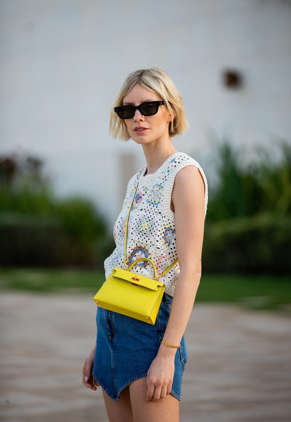 Mulher jovem e branca, de cabelo liso loiro e curto, posa para foto na rua. Ela usa uma blusa de crochê branca, um shorts jeans, uma bolsa amarela pequena da marca Hermès e um óculos escuro. - Metrópoles