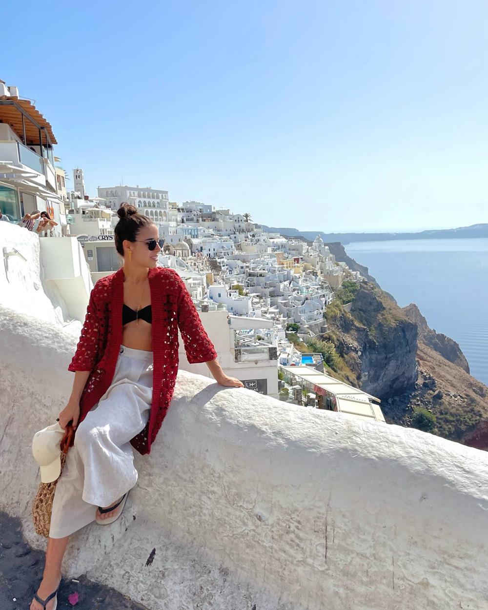 Mulher branca e jovem, de cabelo liso, posa para foto em ponto turístico da Grécia. Usa um top preto de biquíni, uma calça branca e um cardigã de crochê rosa, além de óculos escuros e um chinelo Havaianas preto. - Metrópoles