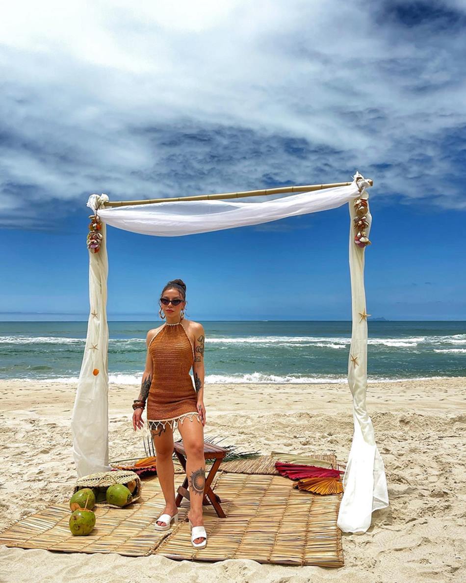 A cantora Priscilla Alcântara, uma mulher branca e jovem, de cabelo liso preto, posa para foto em uma praia. Ela usa um vestido marrom de crochê curto, óculos escuro e um chinelo branco. - Metrópoles