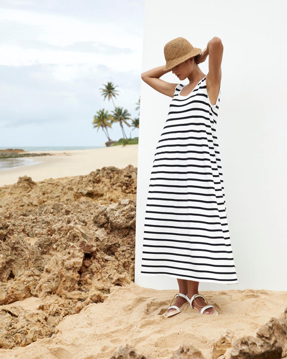 Mulher branca e jovem, de cabelo liso, posa para foto na praia. Ela usa um vestido branco com listras pretas, uma sandália rasteira branca e um chapéu de palha. - Metrópoles