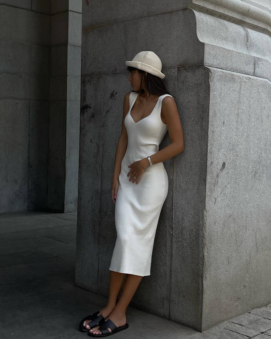 A influenciadora de moda Livia, uma mulher branca, jovem, com cabelo liso castanho, posa para foto encostada em uma pilastra. Ela usa um vestido branco de tricô, uma sandália marrom e um chapéu de crochê branco.