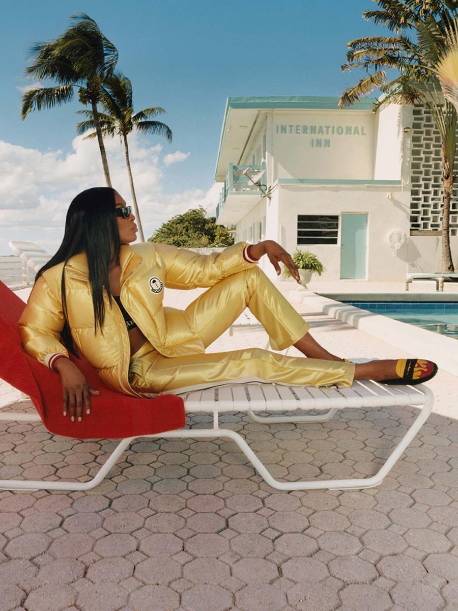 Mulher negra e jovem, de cabelo liso longo, posando para foto sentada em uma cadeira de tomar sol na beira da piscina. Ela usa óculos escuros, uma calça dourada, um casaco puffer dourado e sapatos estilo mocassim preto. Todas as peças são da marca Moncler. - Metrópoles