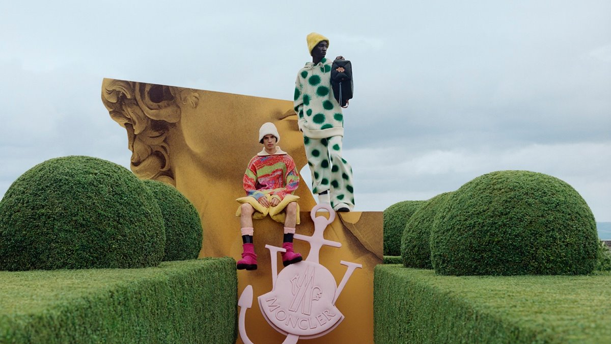 Dois homens jovens, um branco e um negro, posam para campanha publicitária da marca Moncler em um jardim. O primeiro usa uma bermuda amarela e um casaco colorido. O segundo usa um conjunto de moletom e calça brancos com bolas verdes. - Metrópoles