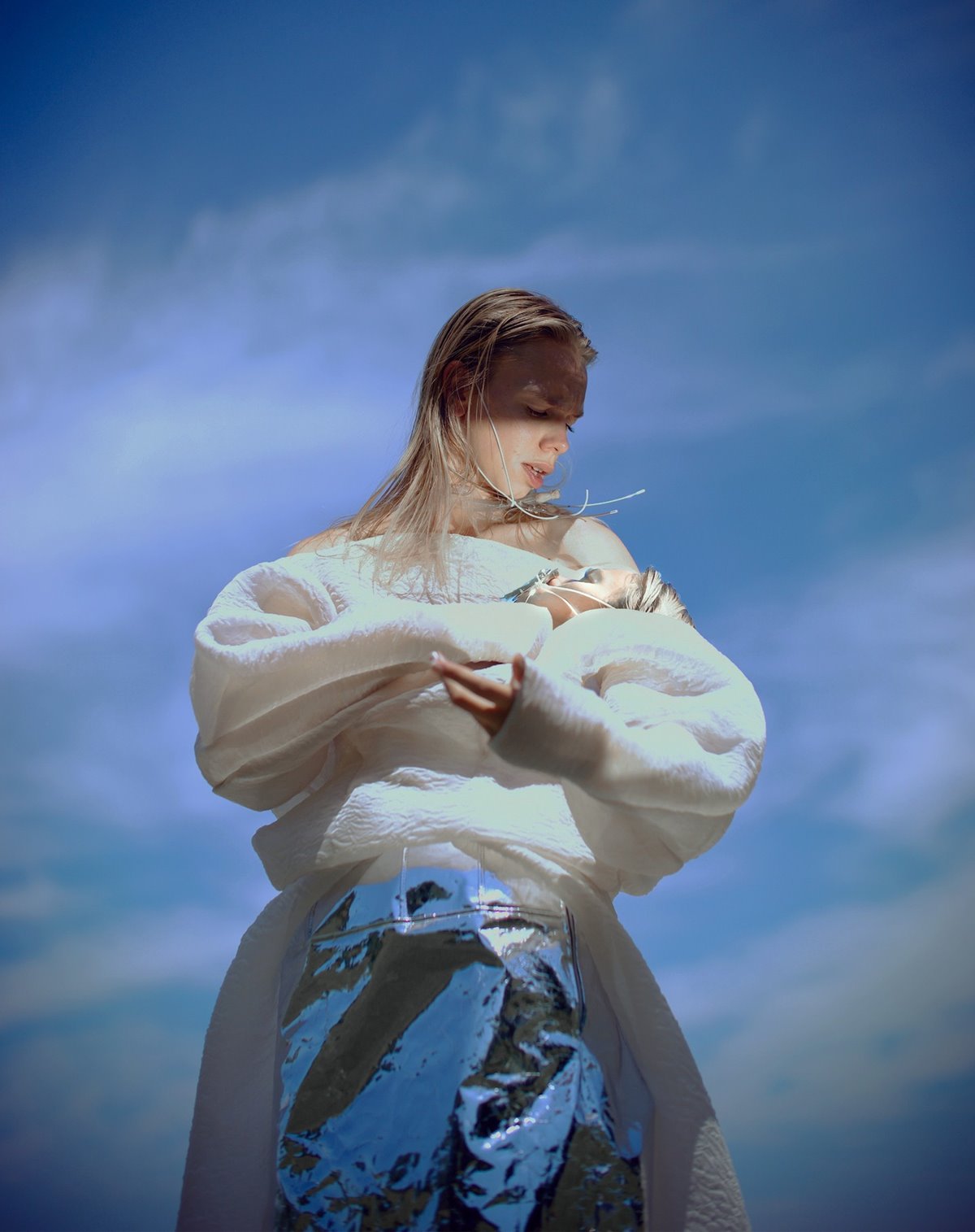 Mulher branca e jovem, de cabelo liso e louro, posa para foto com o céu ao fundo. Ela usa um vestido de manga cumprida branco com um avental-saia prata amarrado na cintura. Segura um bebê na posição de ninar. - Metrópoles