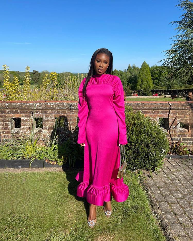 Mulher negra e jovem, de cabelo liso longo, posa para foto em um jardm. Ela usa um vestido longo rosa e uma sandália de salto e uma bolsa, ambos na cor dourada. - Metrópoles