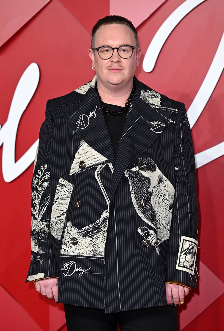 O stylist Harry Lambert em evento de moda em 2022. Ele usa camiseta preta e um blazer, também preto, com gravuras em preto e branco. Ele é um homem branco, jovem, com óculos de grau de armação preta. - Metrópoles