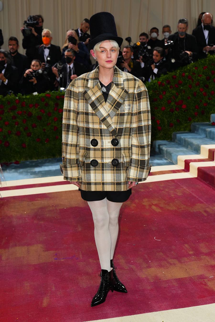 A atriz Emma Corrin, uma mulher jovem e branca, de cabelo liso curto, posa para foto no evento Met Gala em 2022. Ela usa um conjunto de blazer e shorts, ambos pretos, e por cima um casaco xadrez amarelo e branco. - Metrópoles