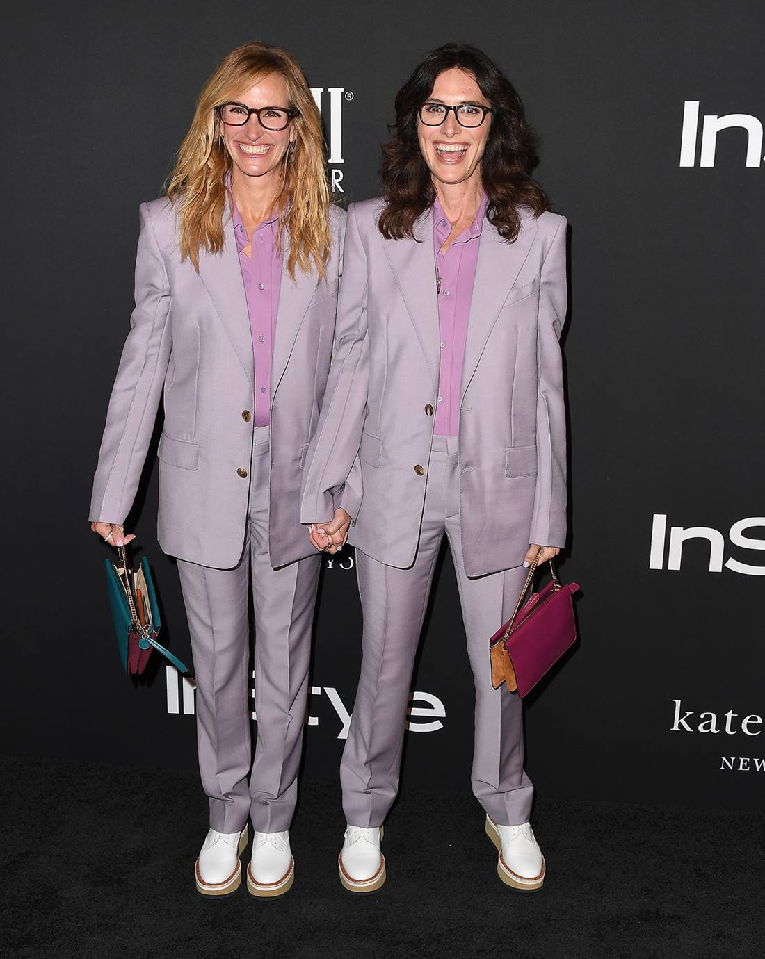 A atriz Julia Roberts e a stylist Elizabeth Stewart vestidas iguais em evento de moda da revista InStyle. Elas usam um conjunto de blazer e calça na cor lilás, uma camiseta roxa e tênis branco. Ambas são mulheres brancas de meia idade com cabelo ondulado, sendo Julia loira e Elizabeth morena. - Metrópoles