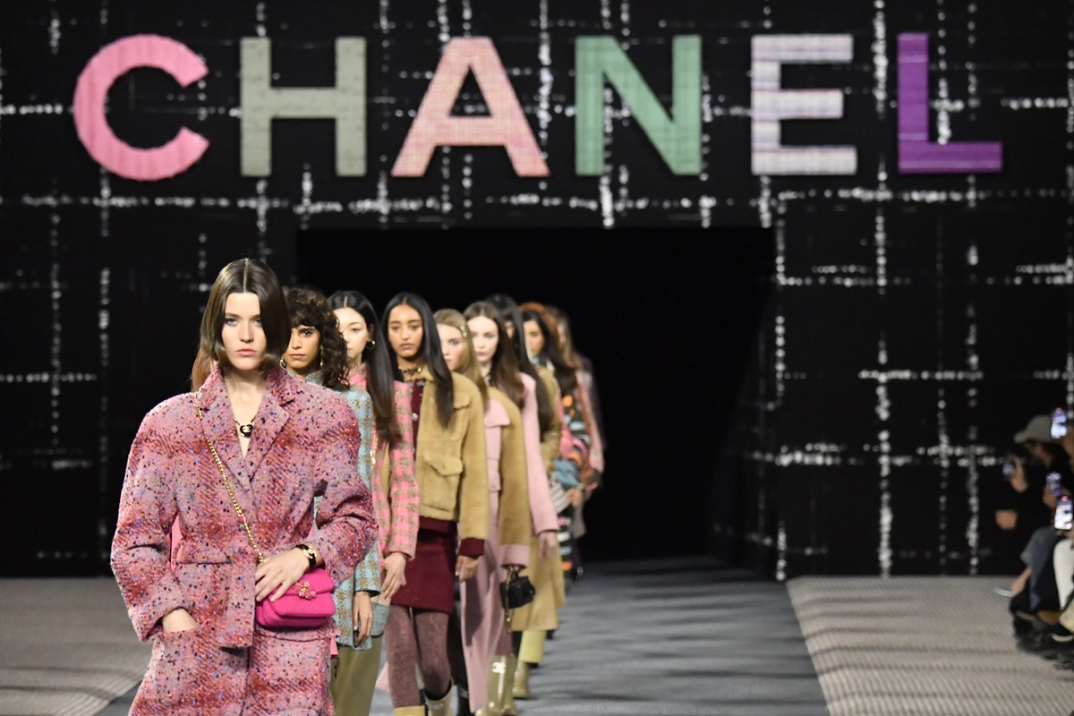 Fila de modelos mulheres no final do desfile da marca francesa Chanel. Ela usam roupas da marca. No fundo, é possível ver um painel de tecido quadriculado com a logo da marca. - Metrópoles