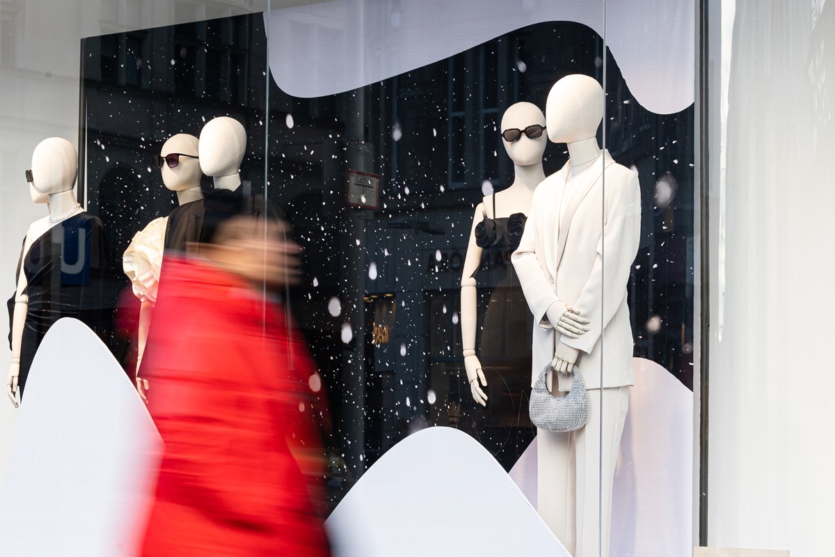 Pessoa passa em frente a uma vitrine de uma loja de moda. Estão dispostos quatro manequins brancos com vestidos e terninhos nas cores preta e branca. Passa em frente à vitrine uma pessoa com casaco vermelho. Ela está borrada e não é possível ver seu rosto. - Metrópoles