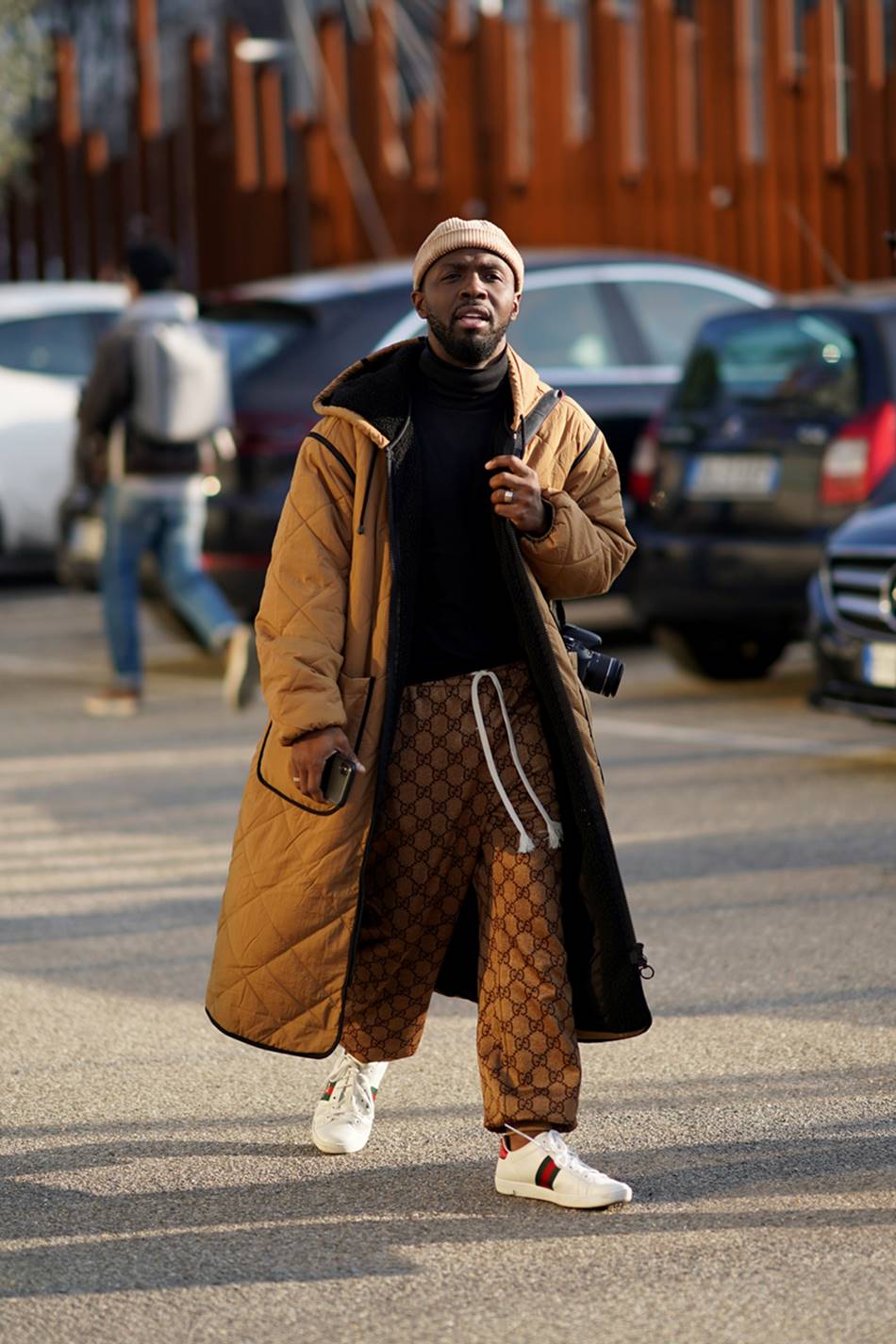 Homem jovem e negro, de cabelo raspado e margem, andando pela rua. Ele usa uma camiseta preta, uma calça marrom a logomarca da grife Gucci, um casaco marrom acolchoado longo e um tênis branco. - Metrópoles