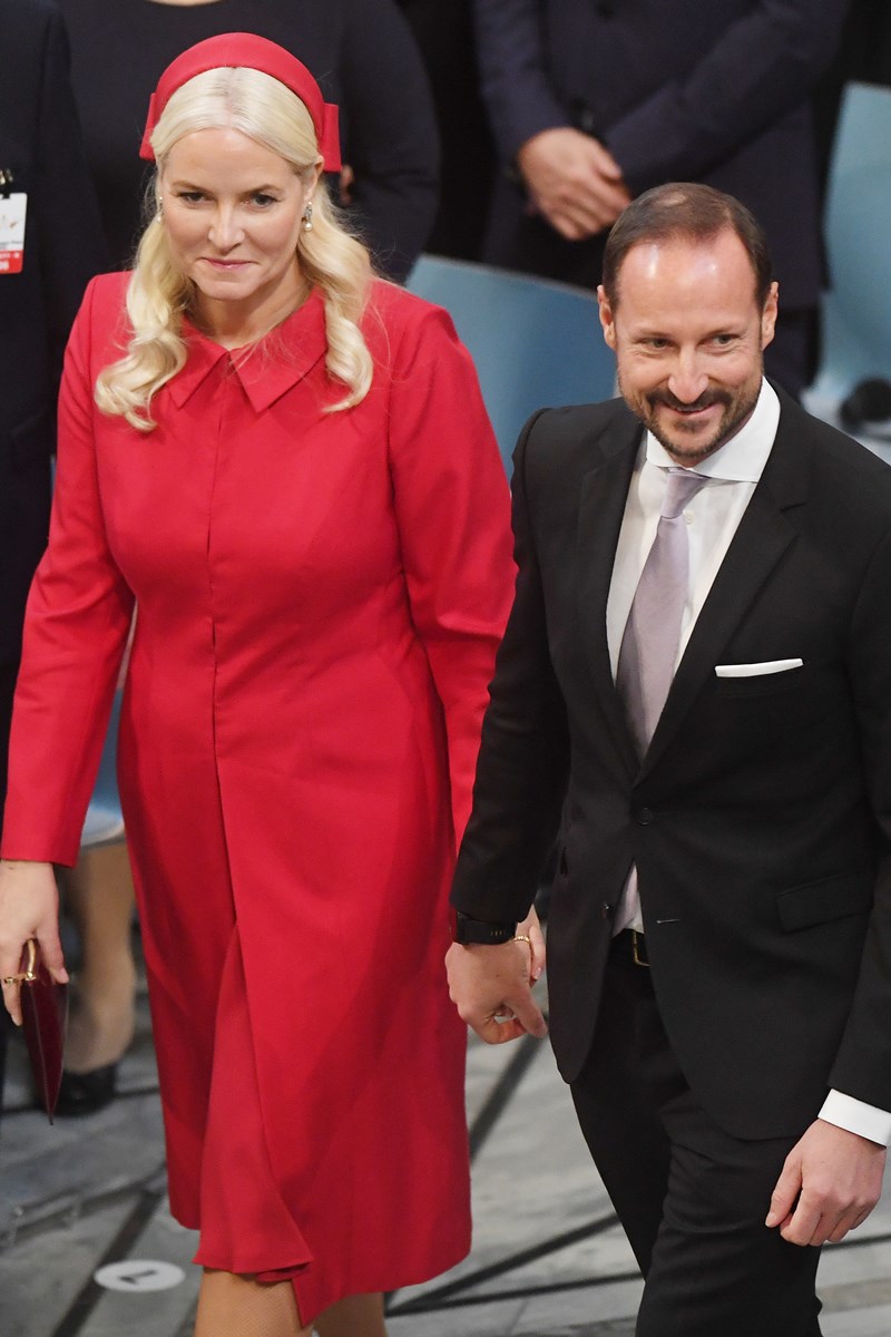 Foto colorida de mulher branca com vestido vermelho de mãos dadas com homem branco e de terno - Metrópoles