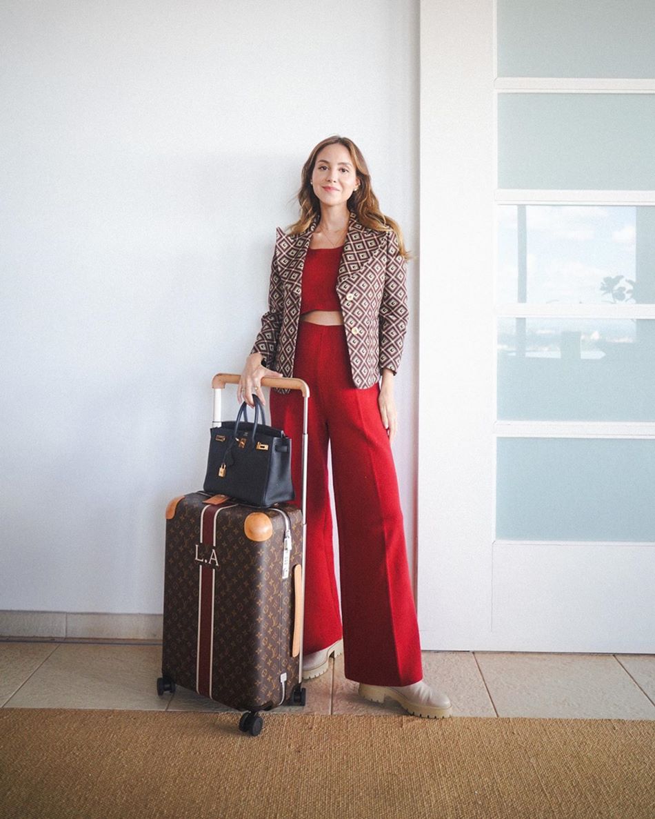 A influenciadora Luisa Accorsi, uma mulher branca e jovem, de cabelo loiro liso, posa para foto em um fundo branco. Ela usa um conjunto de top e calça, ambos vermelho, um blazer marrom e uma bolsa preta. Está segurando ainda uma mala marrom da Louis Vuitton. - Metrópoles