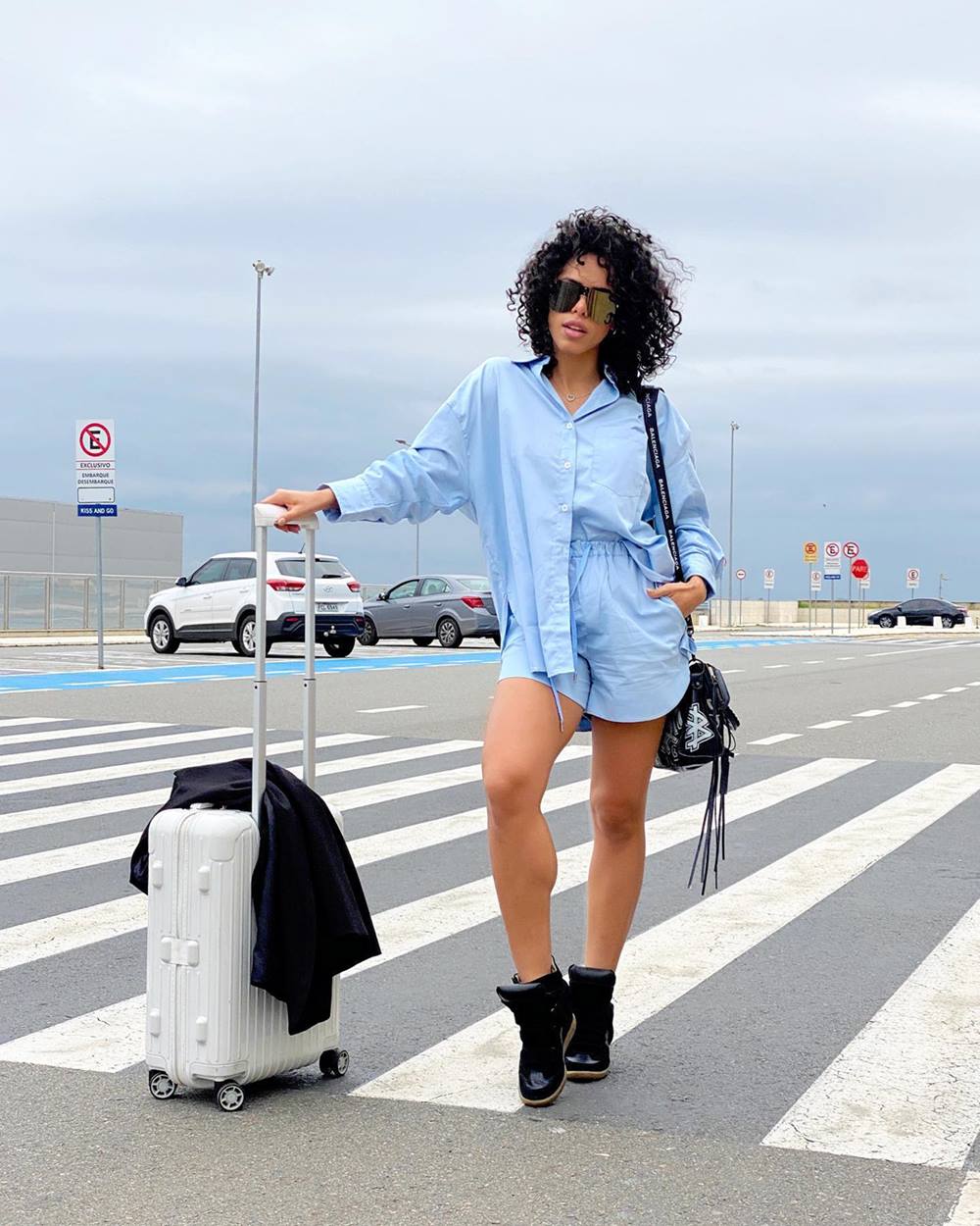 A influenciadora Rayza Nicacio, uma mulher negra e jovem, de cabelo cacheado preto, posa para foto no estacionamento de um aeroporto. Ela usa um conjunto de camisa e shorts, em cor azul claro, um tênis preto, óculos escuros e bolsa de couro preta. Segura uma mala prata. - Metrópoles