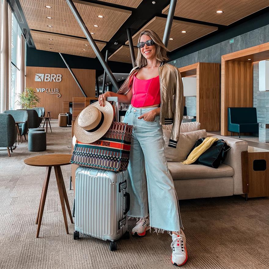 A consultora de moda Helô Drummong, uma mulher branca e jovem, de cabelo liso loiro, posa para foto no aeroporto. Ela usa um top rosa, uma calça jeans, tênis e um casaco marrom por cima dos ombros. Segura uma mala prata, uma bolsa marrom da marca Dior e um chapéu de palha. - Metrópoles