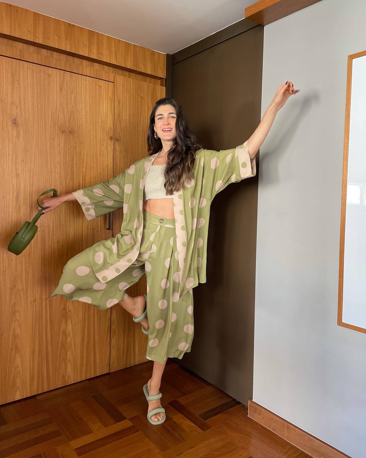 Uma mulher branca e jovem, de cabelo longo ondulado, posa para foto na sala de casa. Ela usa um top branco curto e um conjunto de quimono e calça, ambos verdes com bolas brancas estampadas; além de sandálias rasteiras e uma bolsa verde. - Metrópoles