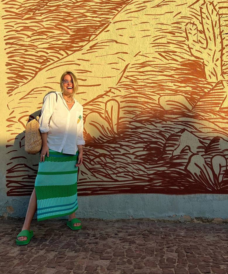 Uma mulher branca e jovem, de cabelo ondulado loiro, posa para foto em frente a um painel amarelo. Ela usa um vestido de crochê verde longo e, por cima, uma camisa de botão branca. Segura uma mochila de palha. - Metrópoles